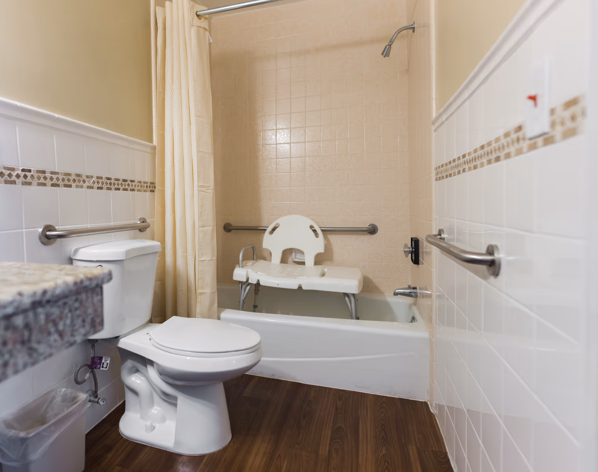 A bathroom with a white toilet, a bathtub with a beige tiled wall, a shower curtain, and a white plastic shower chair inside the tub. The bathroom has grab bars on the walls for accessibility and a granite countertop partially visible on the left.