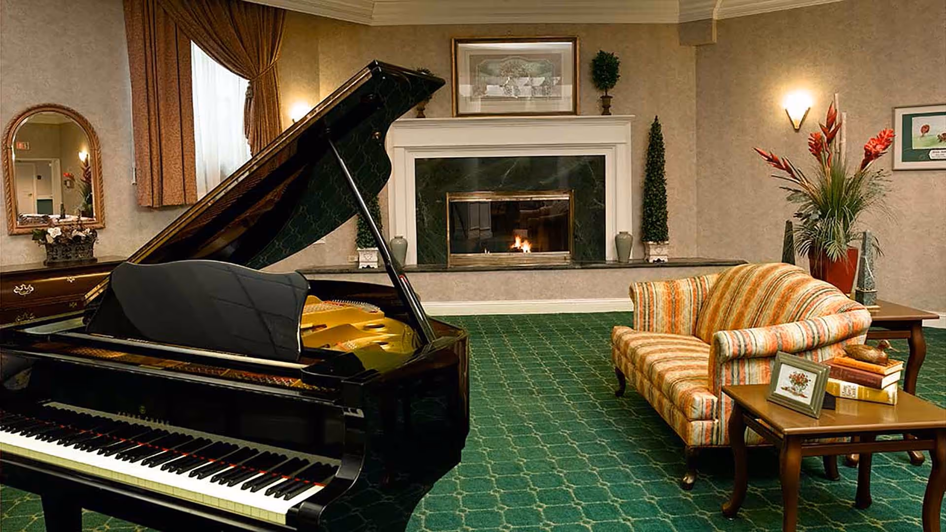 A cozy living room with a grand piano in the foreground, a striped upholstered sofa, a wooden coffee table with books and a framed picture, a fireplace with a fire burning, and decorative plants and artwork on the walls.