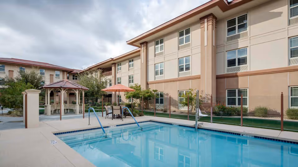 Courtyard swimming pool with chairs, an umbrella and a gazebo in front of a three-story senior living building.
