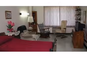 A cozy living room area with a red sofa in the foreground, several armchairs, a wooden coffee table, a TV on a wooden stand, a floor lamp, and a window with sheer curtains. There are decorative items including a vase with pink flowers and framed pictures on the walls.