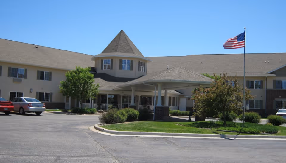 Exterior view of a senior living facility building with a covered entrance, several windows, a few parked cars, green bushes and trees, and an American flag on a flagpole against a clear blue sky.