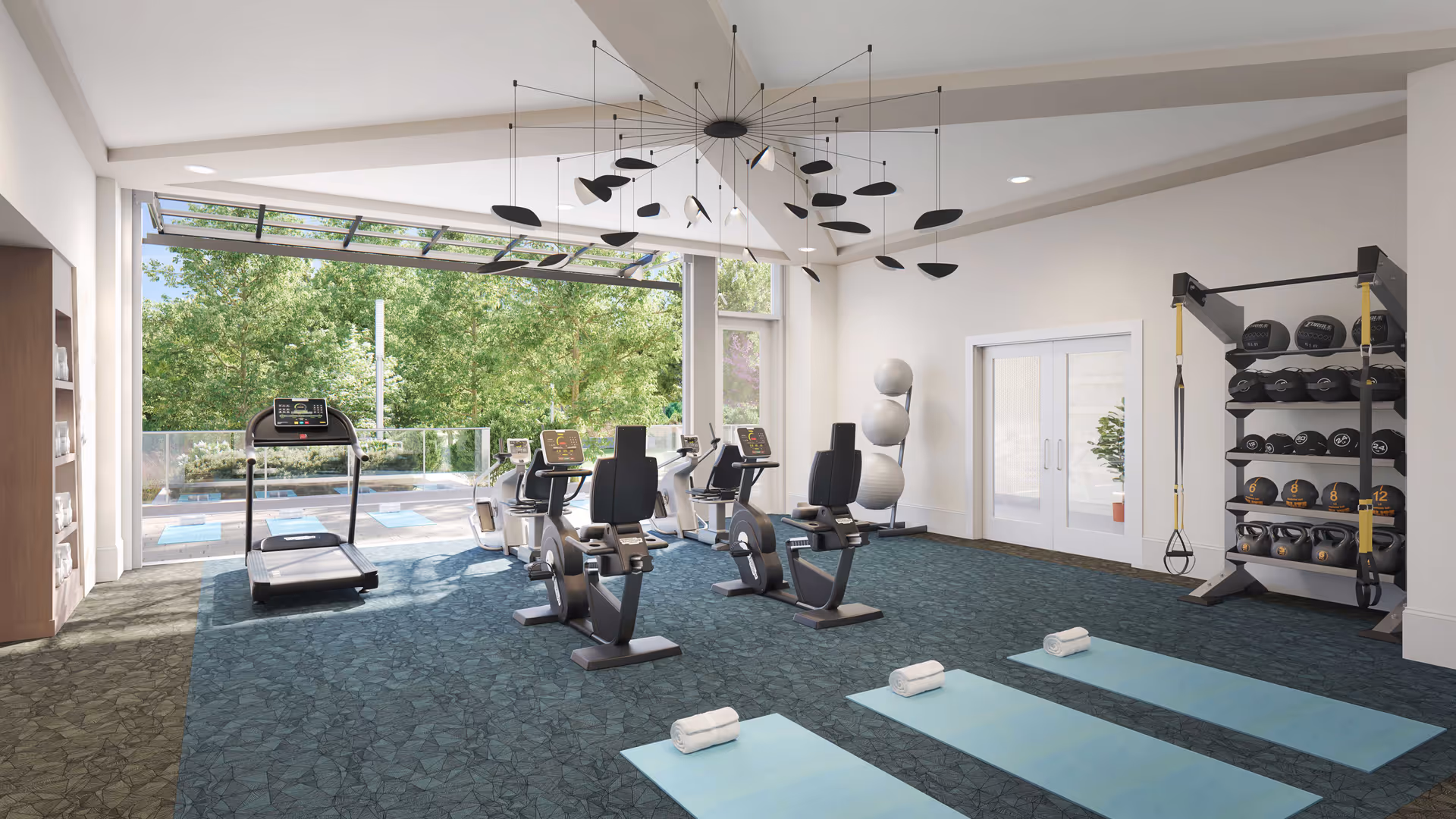 A bright and spacious fitness room with large windows overlooking greenery. The room contains exercise equipment including a treadmill, stationary bikes, yoga mats with towels, a rack of kettlebells and medicine balls, and exercise balls stacked in the corner. The ceiling features a modern black mobile sculpture.