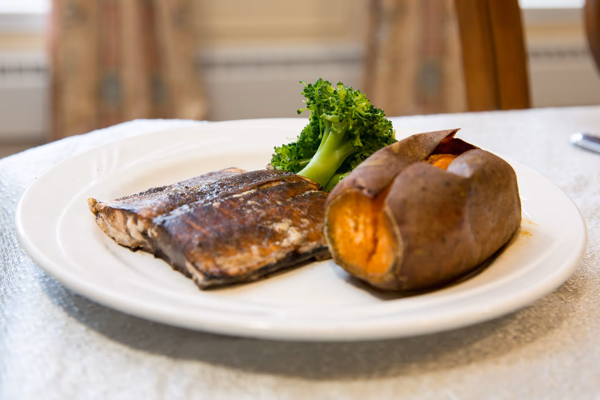 A white plate with a serving of cooked broccoli, a baked sweet potato, and a piece of grilled fish on a table with a white tablecloth.