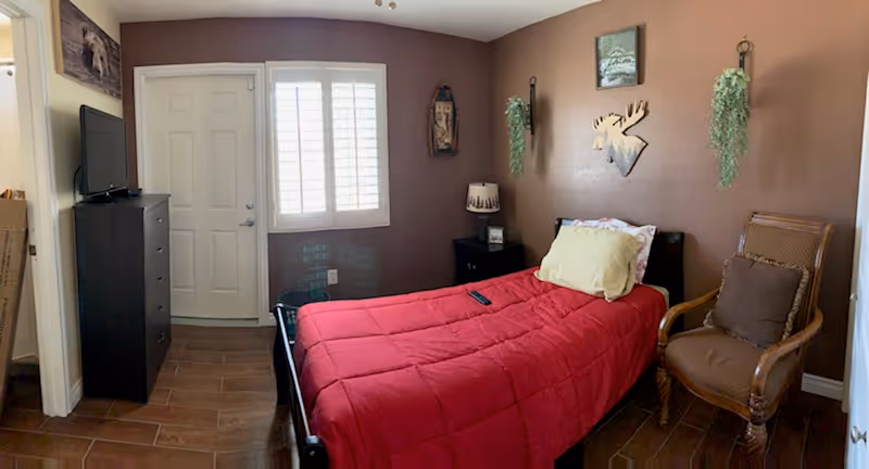 A small bedroom with brown walls and wood-look tile flooring. The room features a single bed with a red comforter and a yellow pillow, a wooden armchair with a brown cushion, a black dresser with a TV on top, a nightstand with a lamp, and wall decorations including a moose head silhouette and hanging greenery. There is a window with white shutters and a door next to it.