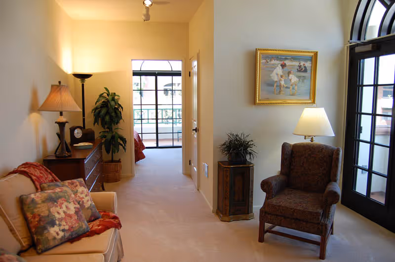 A cozy interior hallway area with a beige carpet and cream-colored walls. On the left side, there is a beige sofa with floral cushions and a red throw, next to a wooden dresser with a lamp, clock, and plant. On the right side, there is a dark upholstered armchair with a floor lamp beside it, a small decorative cabinet with a plant on top, and a framed painting of people at the beach hanging on the wall. Large black-framed windows and a door let in natural light from the outside.