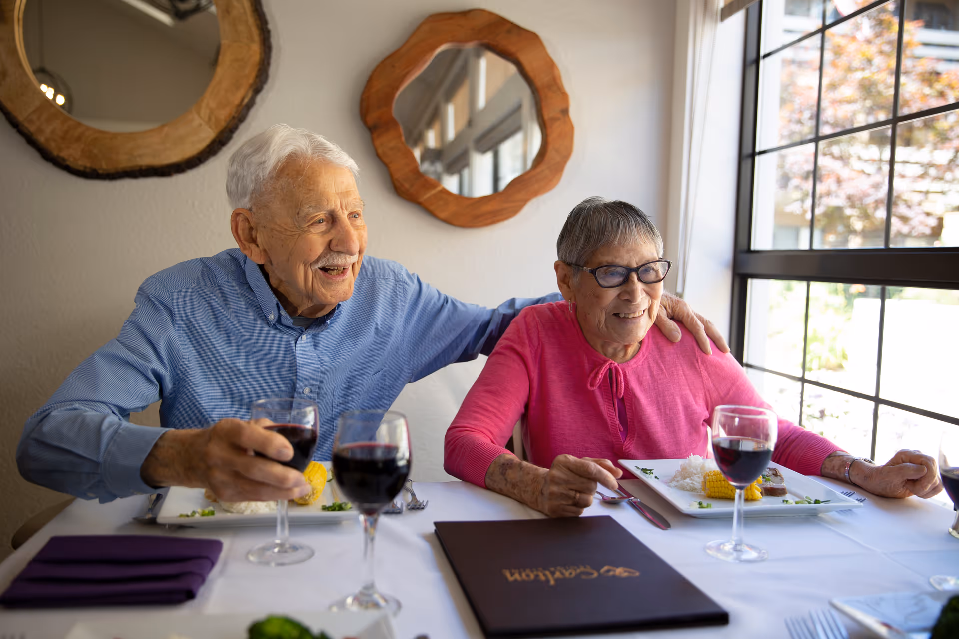 An elderly man and woman sitting at a dining table in a senior living facility, smiling and enjoying a meal together. The man is wearing a blue shirt and holding a glass of red wine, while the woman is wearing a pink top and glasses. There are plates of food and glasses of red wine on the table, with a window letting in natural light in the background.