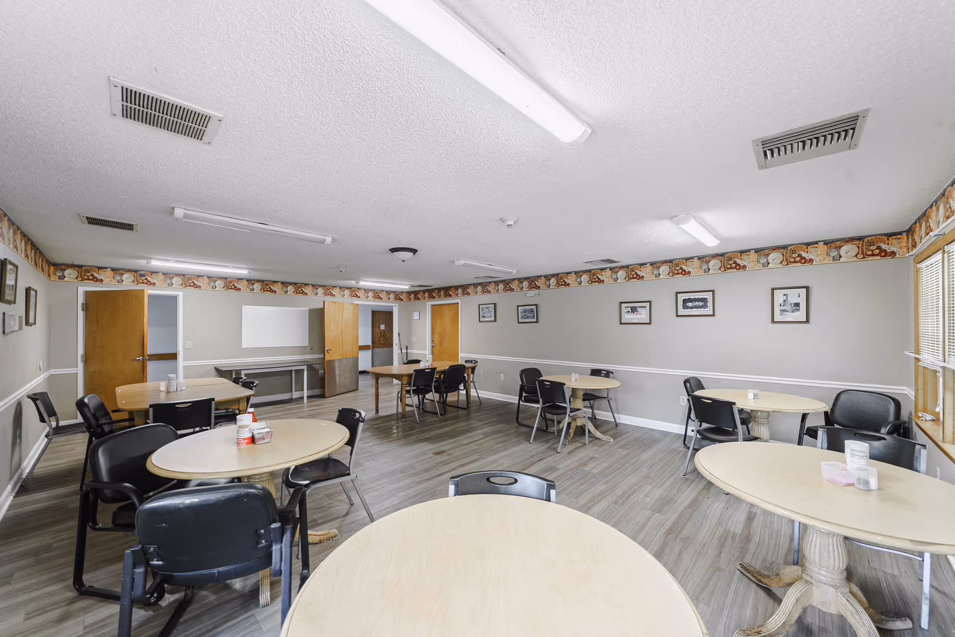 A dining room with several round tables and black chairs arranged around them. The room has light gray walls with a decorative border near the ceiling, wood doors, and large windows letting in natural light. The floor is wood laminate, and fluorescent lights are mounted on the ceiling.