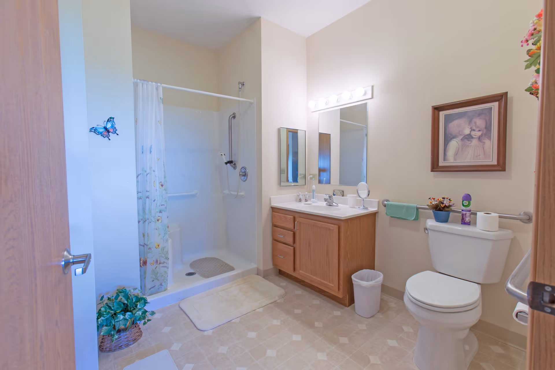 A clean and well-lit bathroom featuring a walk-in shower with a floral shower curtain, a wooden vanity with a sink and mirror above it, a toilet with a small plant and air freshener on the tank, a towel rack with a green towel, a small trash bin, and a framed picture on the wall.