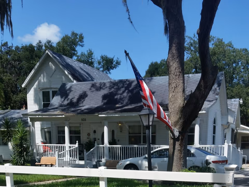 A white wooden house with a porch and white railing, featuring two benches on the porch. An American flag is mounted on a pole attached to a tree in front of the house. A silver car is parked beside the house, and there is a white fence in the foreground. The sky is clear and blue with some trees in the background.