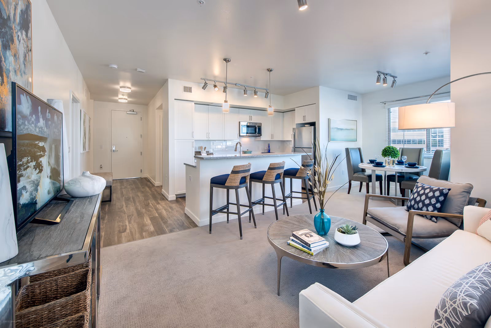 A modern open-concept living area featuring a living room with a white sofa, armchair, and round coffee table with decorative items. Adjacent to the living room is a kitchen with white cabinetry, a breakfast bar with three stools, and stainless steel appliances. In the background, there is a dining area with a round table set for four and a large window letting in natural light.