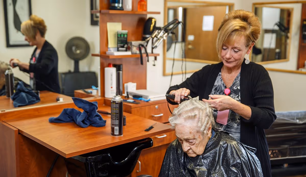 A hairstylist is cutting the hair of an elderly woman seated in a salon chair. The setting appears to be a hair salon with wooden counters, mirrors, and hair care products visible. The elderly woman is wearing a black salon cape, and the hairstylist is focused on her work.
