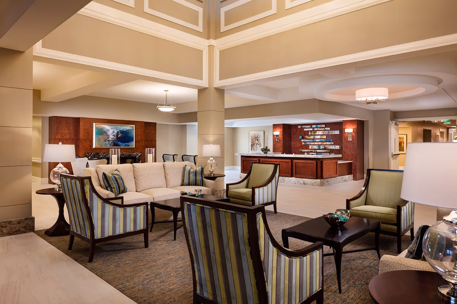 A spacious and well-lit living room area in a senior living facility featuring a beige sectional sofa with striped accent pillows, several striped armchairs, dark wood coffee tables, and side tables with lamps. The room has a high ceiling with decorative molding and a chandelier. In the background, there is a reception desk with a decorative wall and artwork.