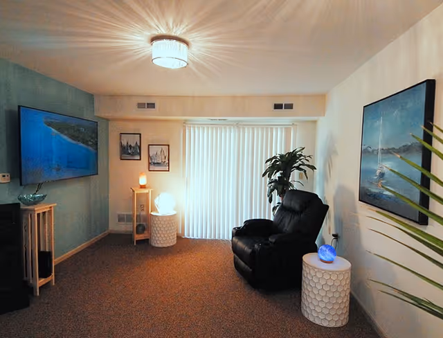 A cozy living room featuring a black recliner, wall-mounted TV, artwork, side tables with lamps, and vertical blinds over sliding doors.