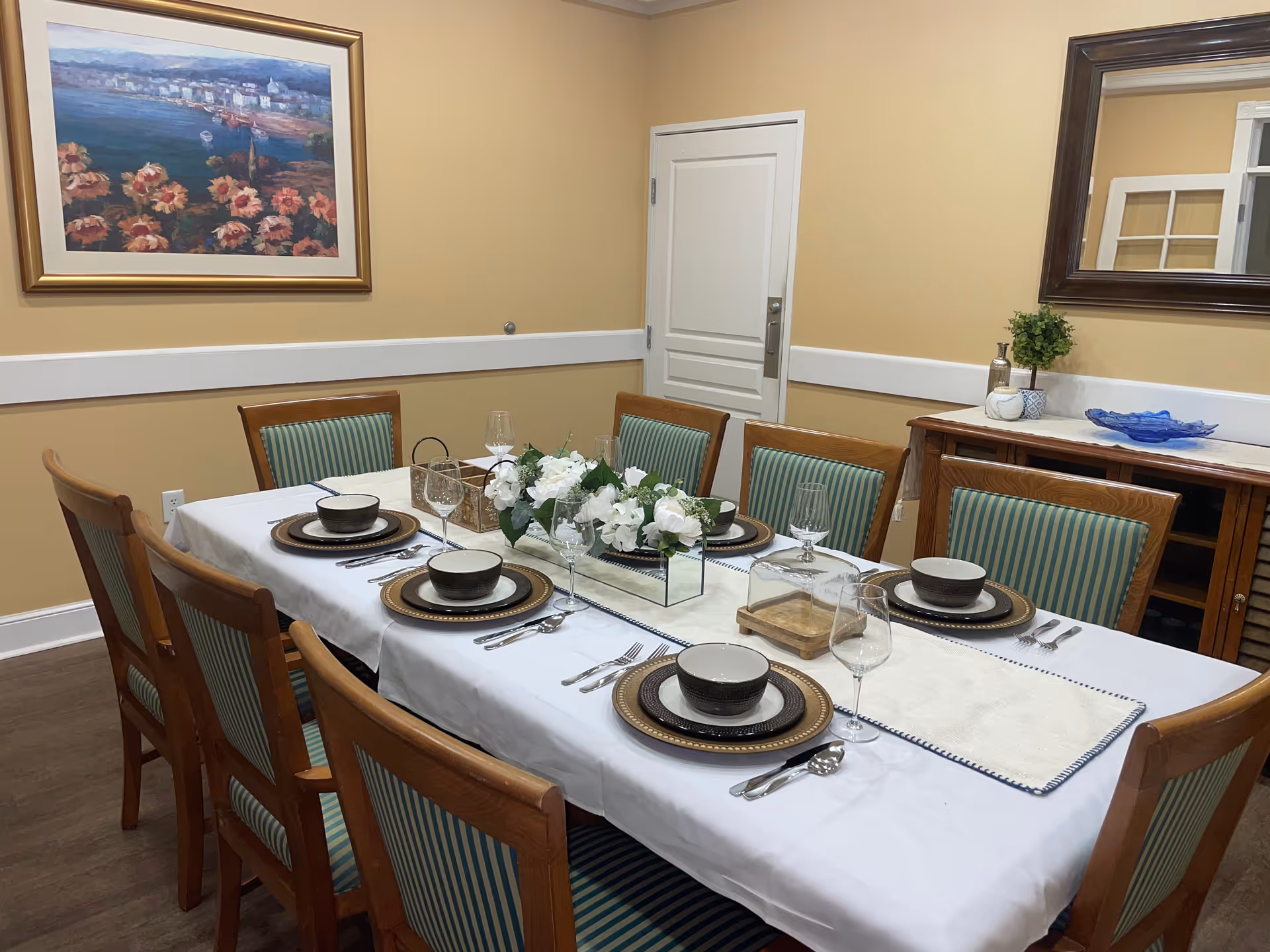 A dining room with a rectangular table set for six people. The table is covered with a white tablecloth and has plates, bowls, wine glasses, and silverware neatly arranged. A centerpiece of white flowers in a rectangular glass vase is in the middle of the table. The room has beige walls, a framed painting on one wall, a large mirror on another, and a wooden sideboard with decorative items.