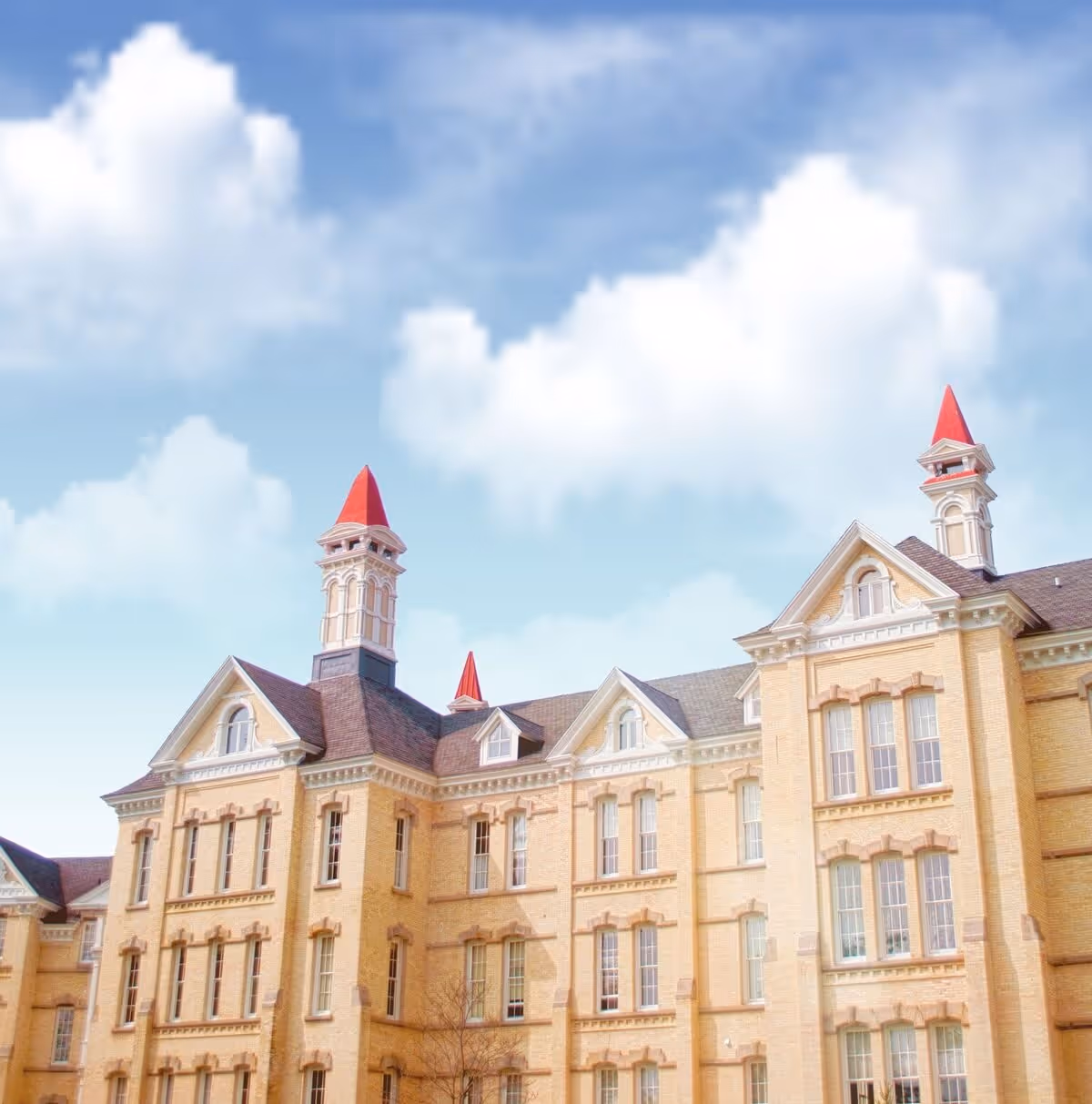 Exterior view of a large historic building with light yellow brick walls and multiple windows. The building features three prominent towers with red pointed roofs under a partly cloudy blue sky.