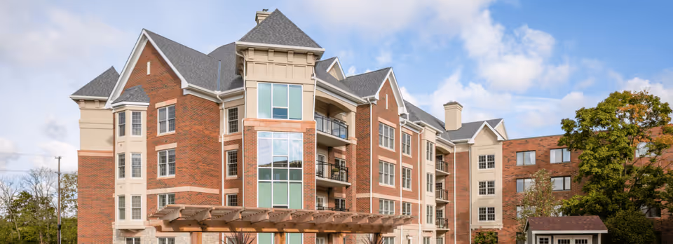 Exterior view of a multi-story senior living facility building with red brick and beige accents, multiple windows, balconies, and a wooden pergola in front. The sky is partly cloudy and there are trees around the building.