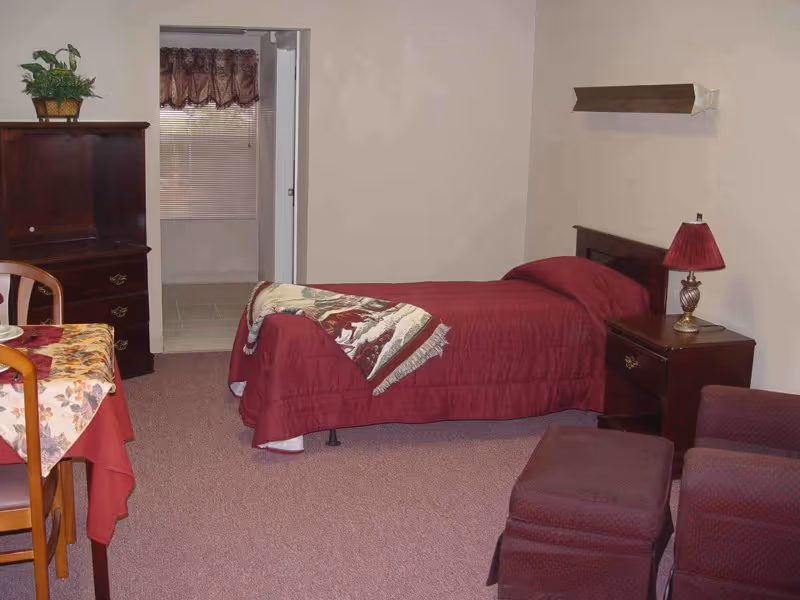 A small assisted-living bedroom with a single bed, nightstand, dresser and a seating/dining area.