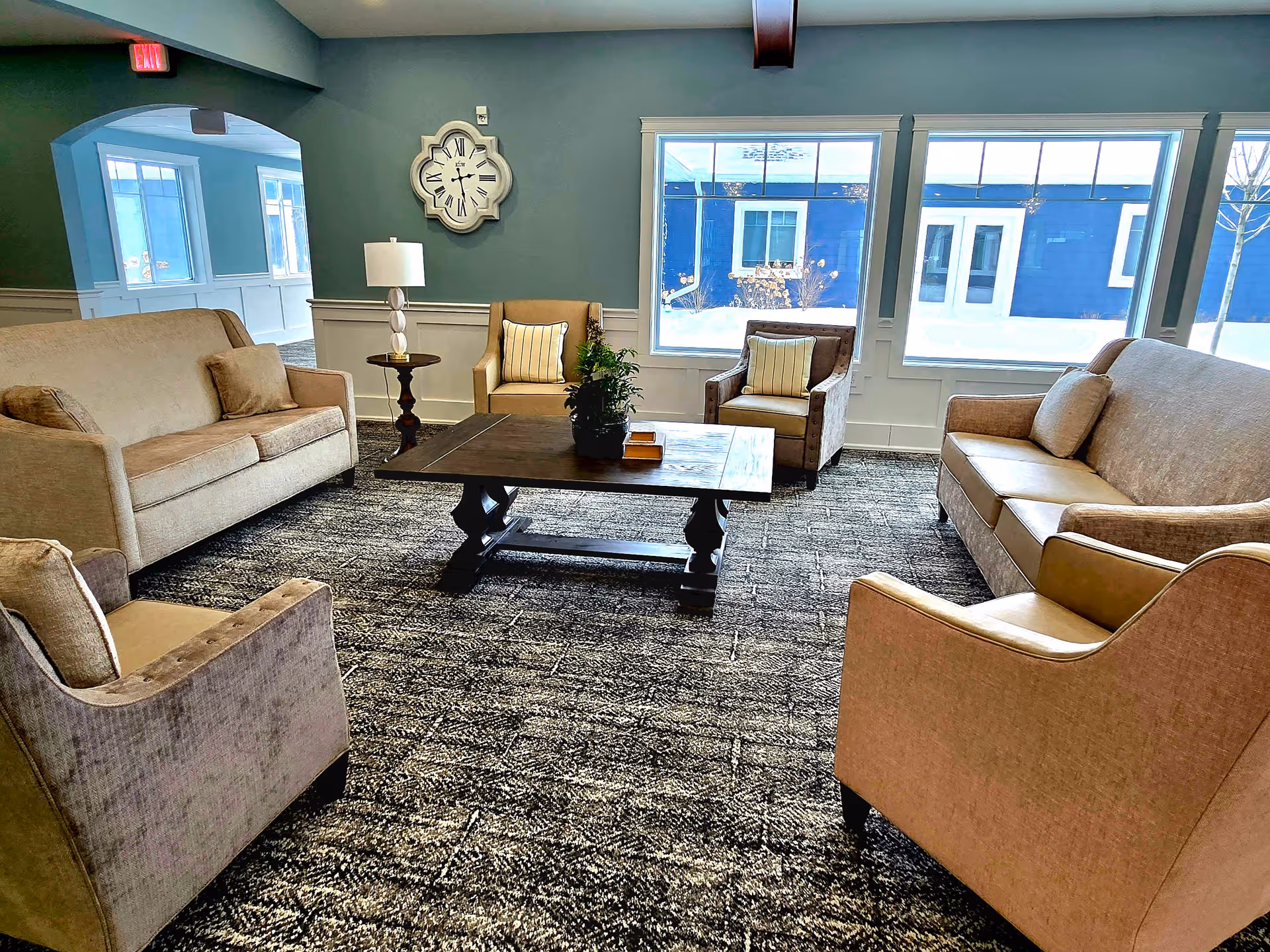 A cozy living room area with two beige sofas, two armchairs, a wooden coffee table with a plant and books on it, a side table with a lamp, a large wall clock, and large windows showing a snowy outdoor scene.