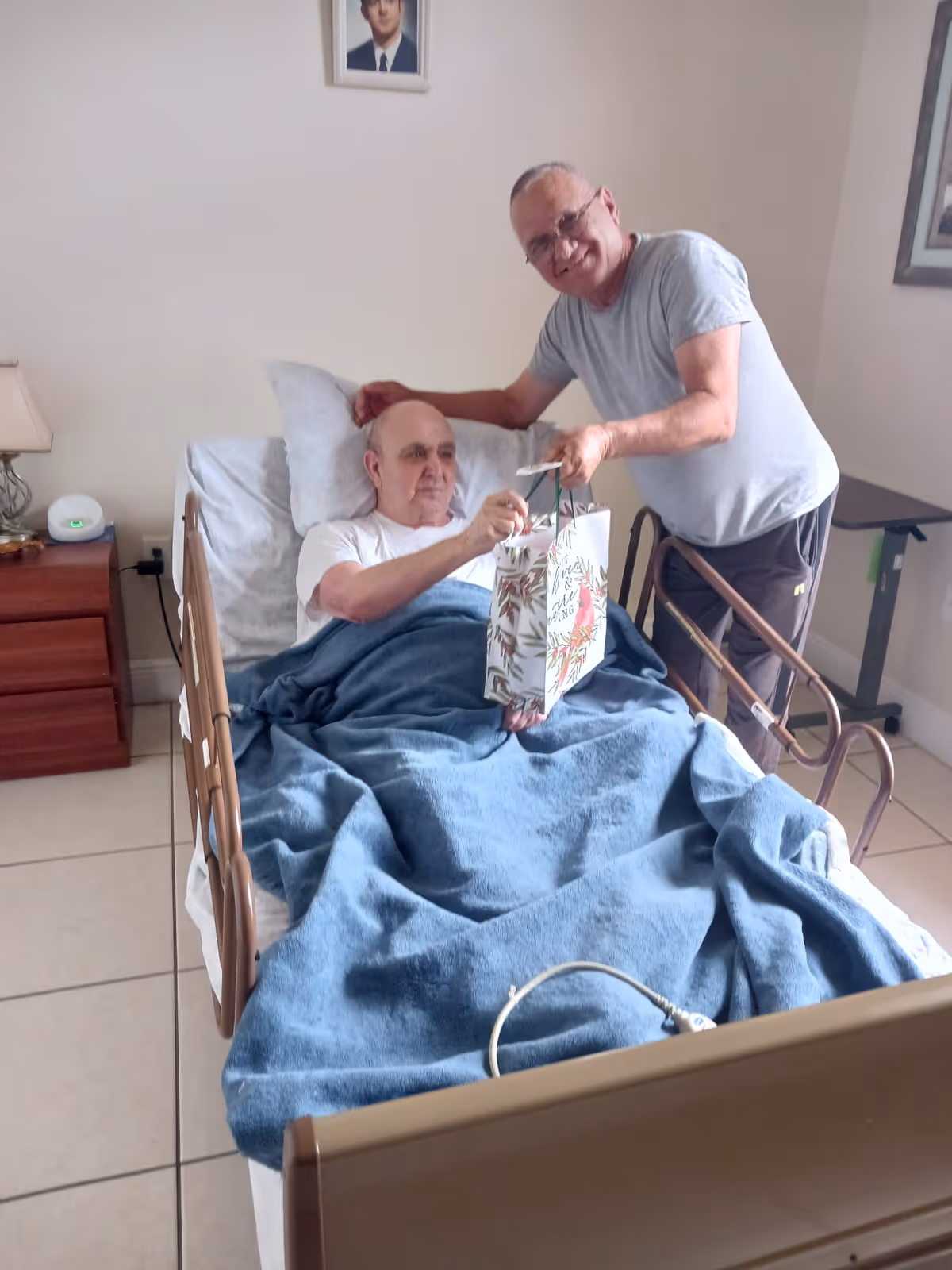An elderly man lying in a hospital-style bed covered with a blue blanket, receiving a gift bag from another smiling elderly man standing beside the bed in a room with tiled floor and simple furnishings.