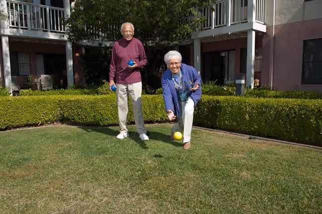 Two elderly individuals playing bocce ball on a grassy lawn area outside a residential building with balconies and trimmed hedges.