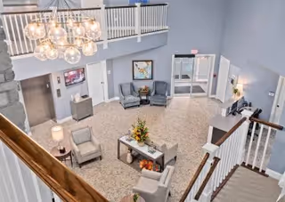 View from an upper floor overlooking a spacious and well-lit lobby area with light gray walls, a modern chandelier with multiple globe lights, a stone fireplace on the left, seating area with armchairs and a small table with flowers, a reception desk on the right, and double glass entrance doors at the far end.