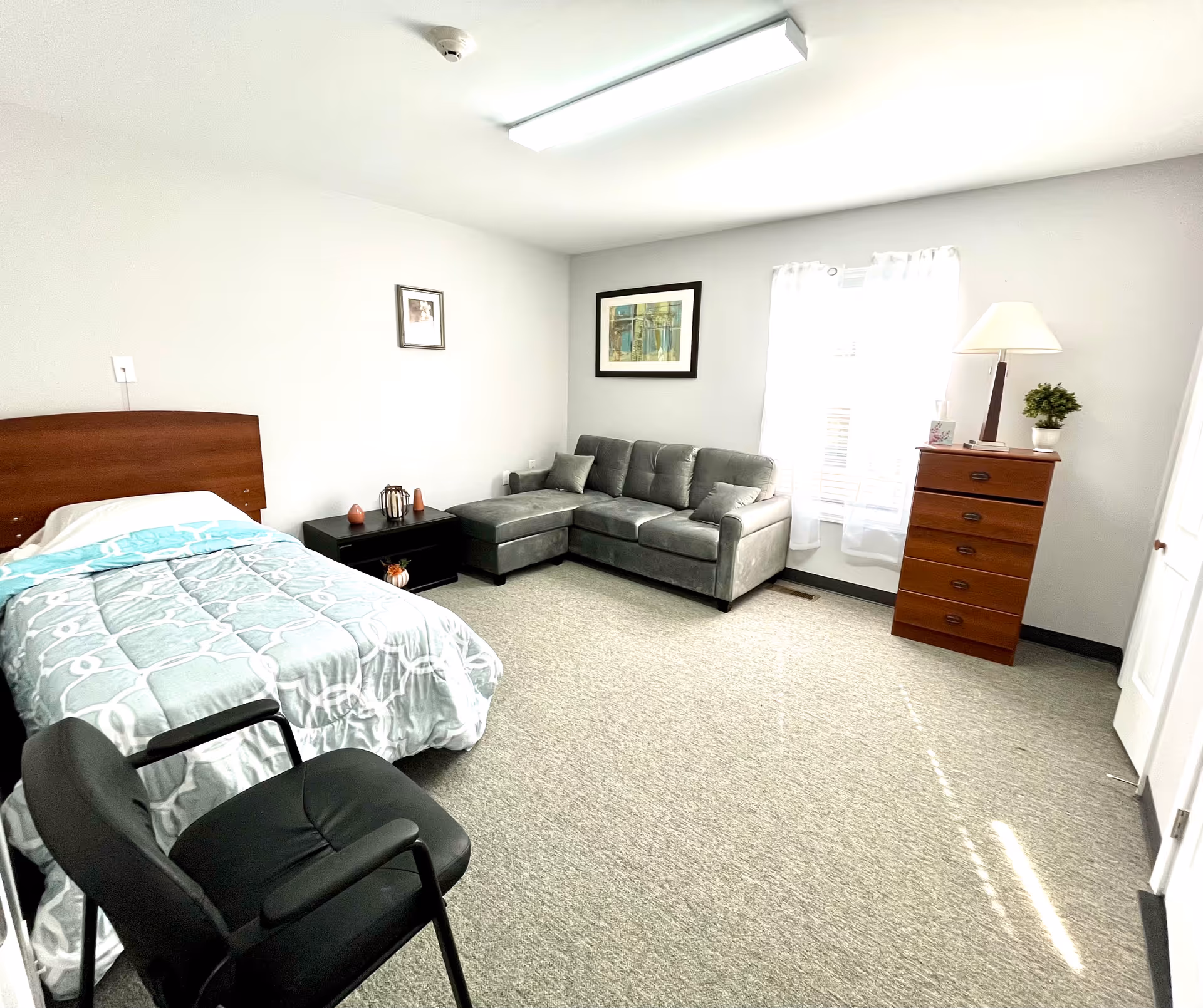 Bedroom in a senior living facility with bed, sofa, dresser, and window.