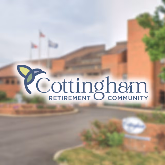 Exterior view of a brick building with a driveway and landscaped greenery, featuring three flagpoles with flags in front of the building. The image has a logo overlay that reads 'Cottingham Retirement Community'.