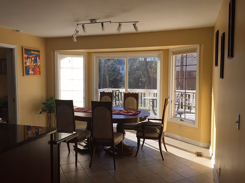 A dining area with a round wooden table and six chairs placed around it. The room has large windows and a sliding glass door that lets in natural light and offers a view of an outdoor patio with additional seating. The walls are painted yellow, and there is a track light fixture on the ceiling. A small plant and a colorful painting are visible on the left side.