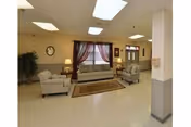 Community lounge with several sofas and armchairs arranged around a rug, lamps, and a window with curtains.