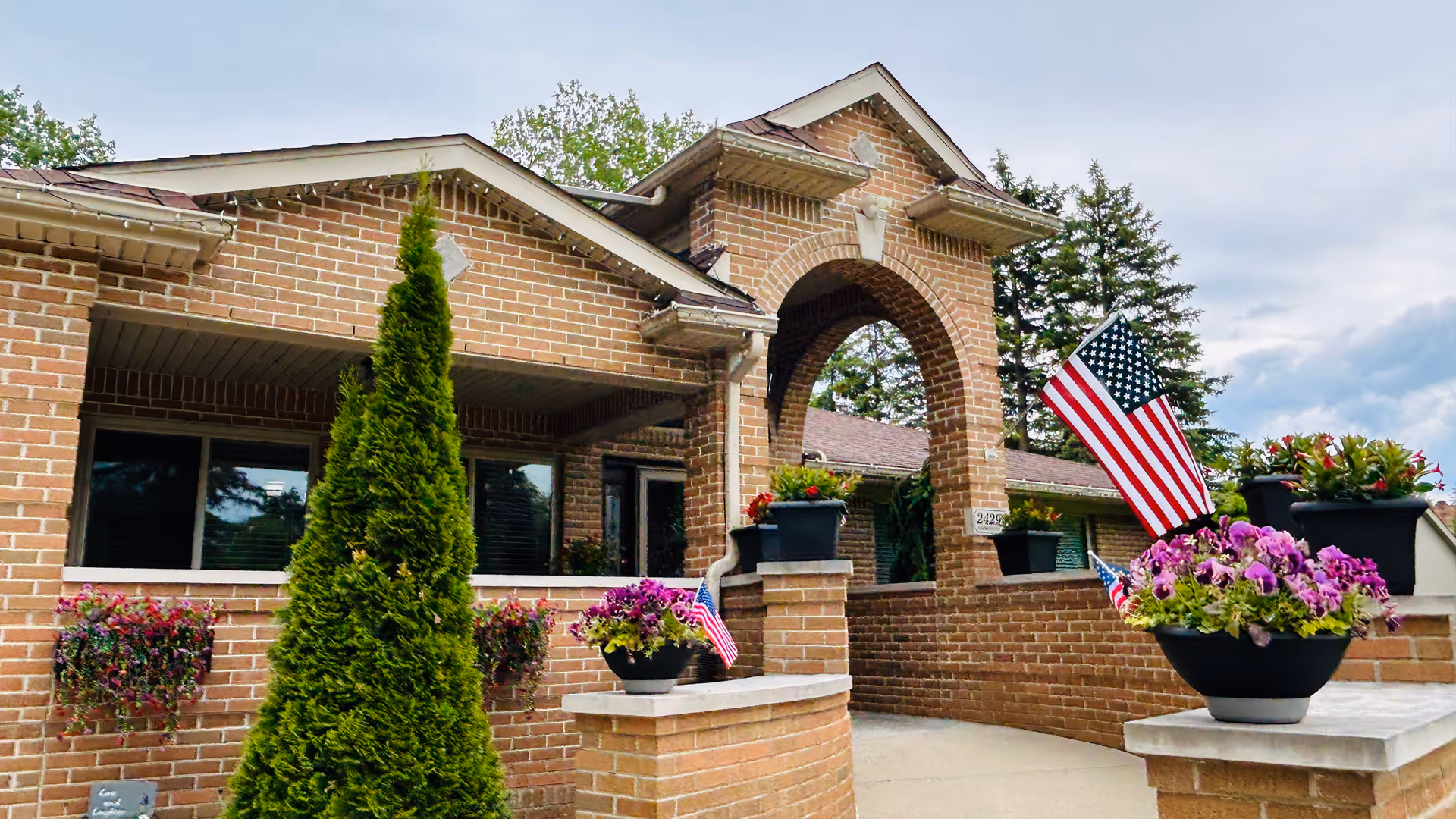 Exterior view of a brick building with an arched entrance, decorated with flower pots containing purple flowers and American flags. There are green trees in the background under a cloudy sky.