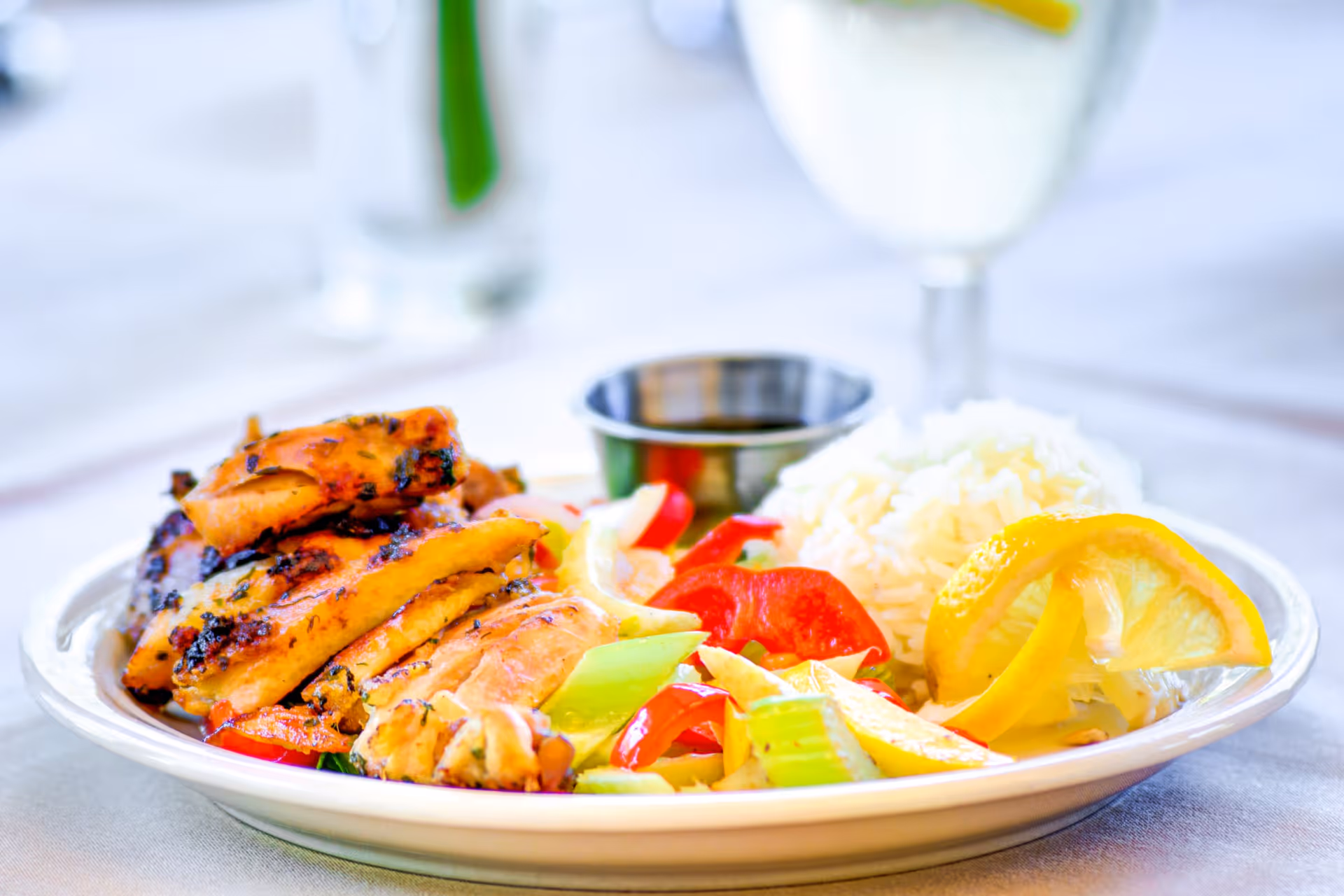 A plate of grilled chicken served with white rice, mixed vegetables including red bell peppers and celery, and lemon wedges. A small metal cup of sauce is also on the plate. The background shows a blurred glass of water and a white tablecloth.