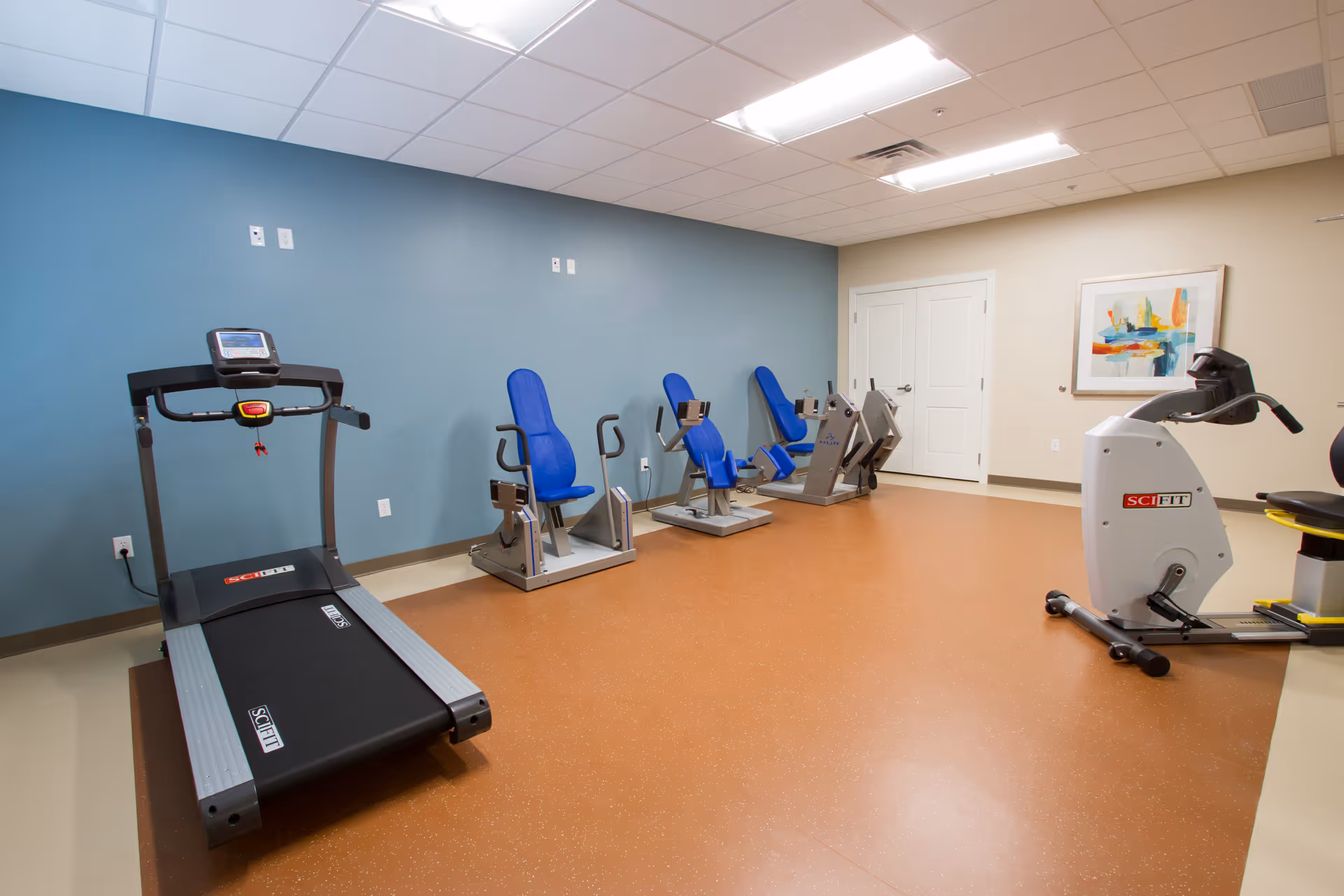 A therapy or exercise room with a treadmill, three blue exercise chairs with attached equipment, and a stationary bike. The room has a blue accent wall, beige walls, a brown floor, and a colorful abstract painting on one wall.