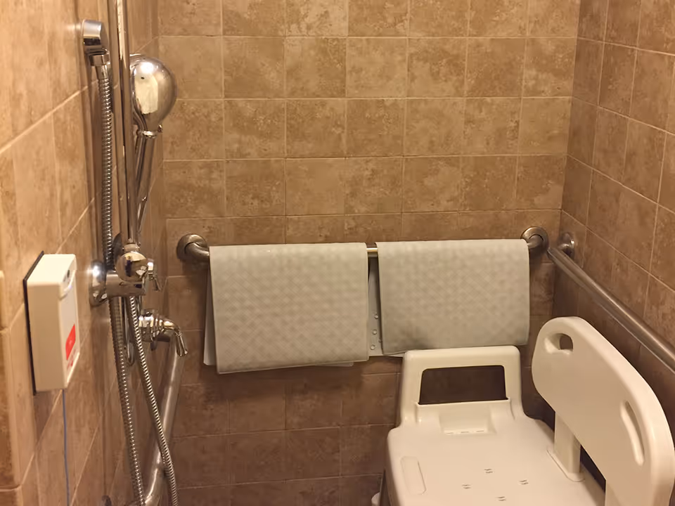 A tiled shower area with beige tiles, featuring a handheld showerhead attached to a vertical bar, two folded white towels hanging on a horizontal grab bar, and a white plastic shower chair with armrests. There is also a call button on the left wall.