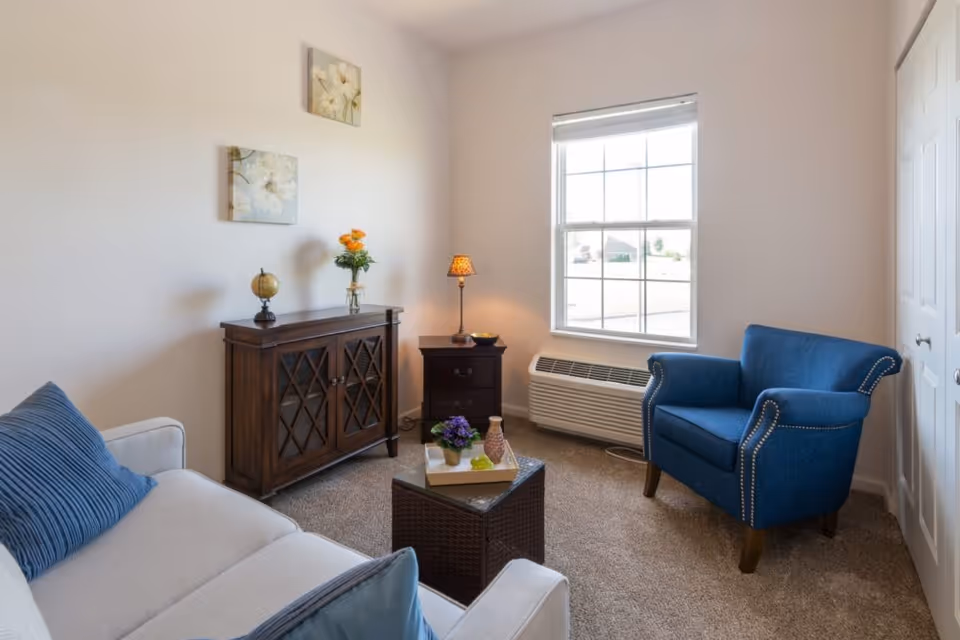 A cozy living room with a beige sofa adorned with blue pillows, a blue armchair with nailhead trim, a dark wooden cabinet with decorative items including a small globe and a vase with orange flowers, a small dark wooden side table with a lamp, and a window letting in natural light. The walls are light-colored and two floral paintings hang above the cabinet.