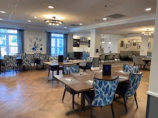 A spacious dining room with several tables and chairs arranged neatly. The chairs have blue and white patterned upholstery. The room features large windows allowing natural light, light-colored walls, and ceiling lights. There are decorative wall art pieces and a fireplace in the background.