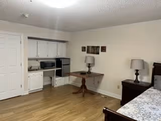 A senior living facility room with a wooden floor, a bed with a patterned bedspread on the right, a wooden nightstand with a lamp, a small wooden table with a lamp against the wall, and a kitchenette area with white cabinets, a microwave, and a small oven. The walls are light-colored and there is a closed white door on the left.