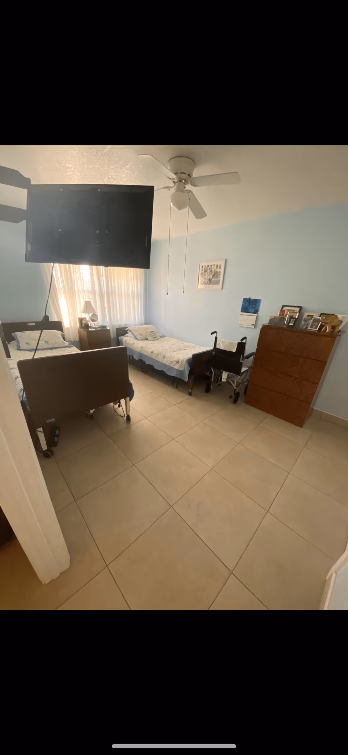 Shared bedroom with two single beds, a dresser, a wheelchair, a wall-mounted TV and a ceiling fan.
