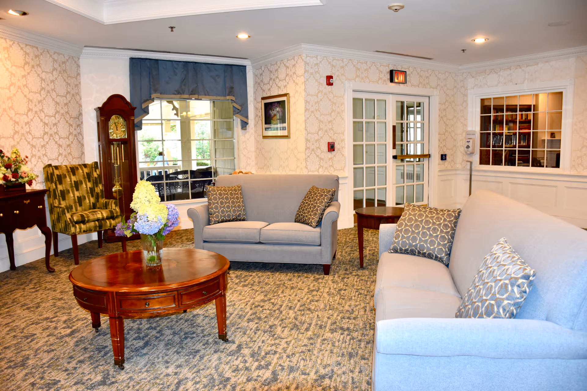 A cozy living room area in Grove Manor Estates featuring two light gray sofas with patterned throw pillows, a round wooden coffee table with a vase of flowers, a green patterned armchair, a grandfather clock, and a side table with a floral arrangement. The room has patterned wallpaper, carpeted floor, and a window with blue valance curtains. French doors and a window looking into another room are visible in the background.