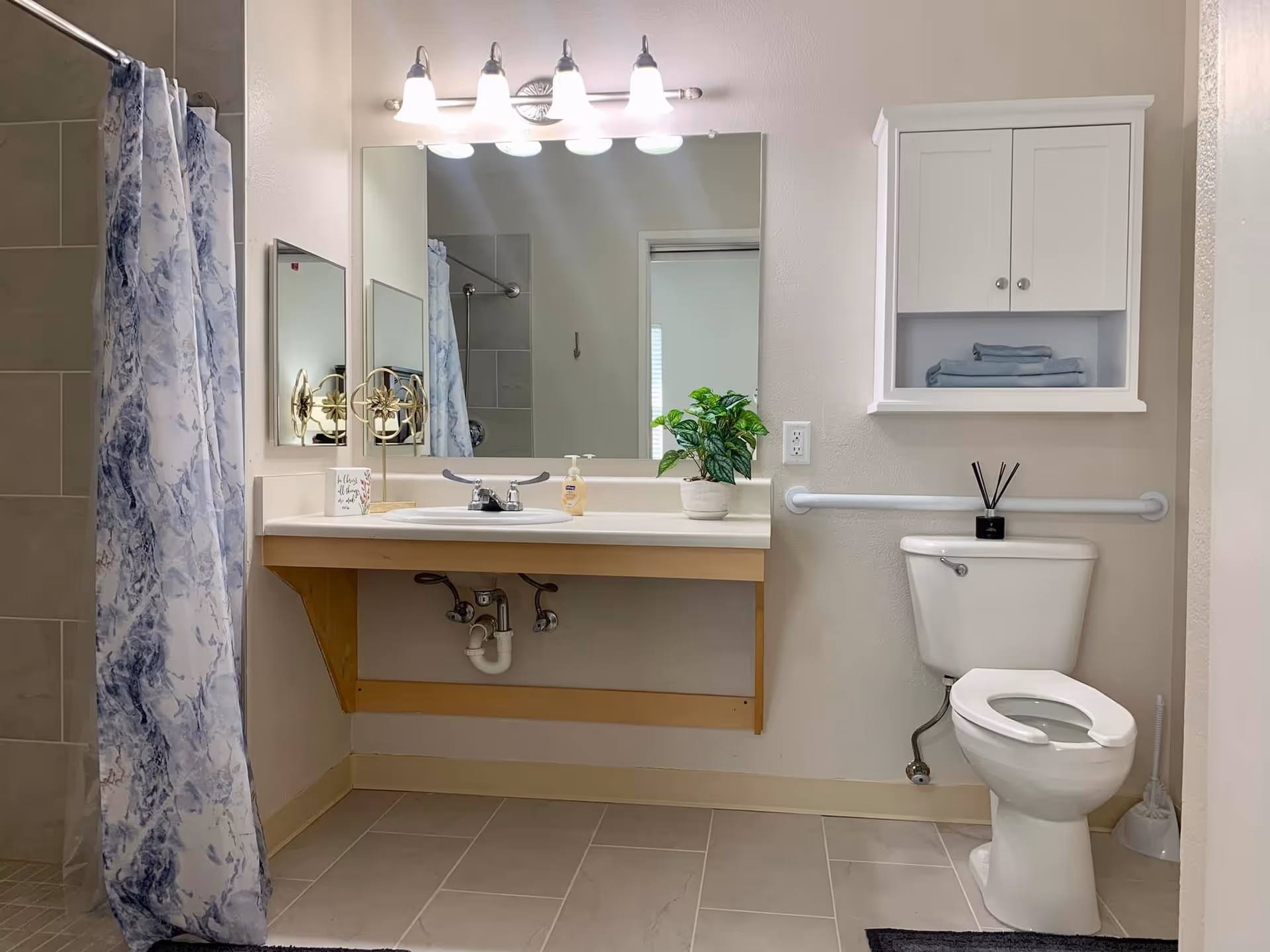 A clean and accessible bathroom featuring a white toilet with a grab bar behind it, a white wall-mounted cabinet with folded towels, a sink with a large mirror above it, a soap dispenser, a small plant, and a shower area with a blue and white patterned curtain.