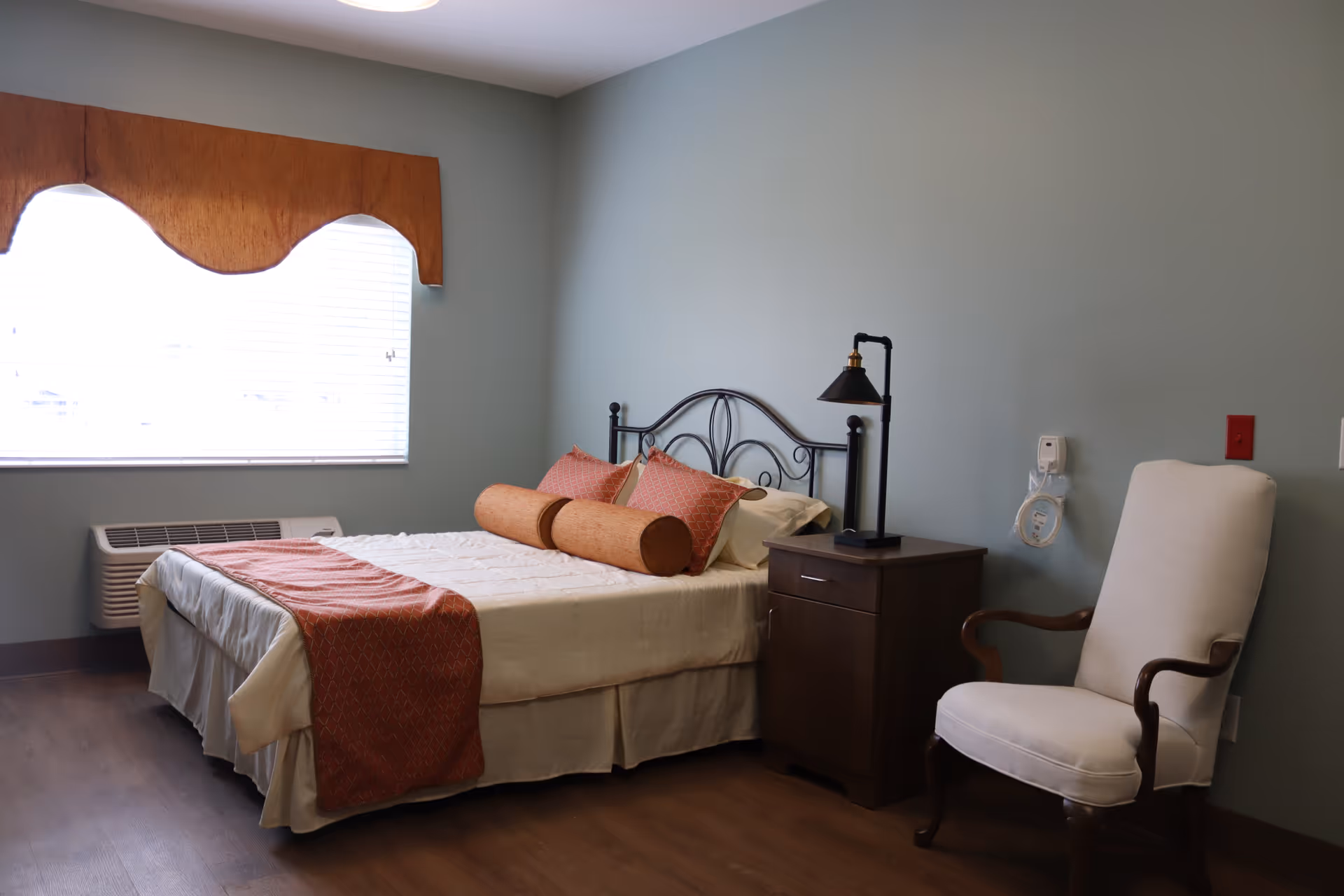 A bedroom with a bed featuring a metal headboard, white bedding, and orange decorative pillows and throw. Next to the bed is a dark wooden nightstand with a black lamp. A white upholstered armchair with wooden armrests is positioned to the right. A window with white blinds and a wooden valance is on the left wall, and there is a wall-mounted air conditioning unit below the window.