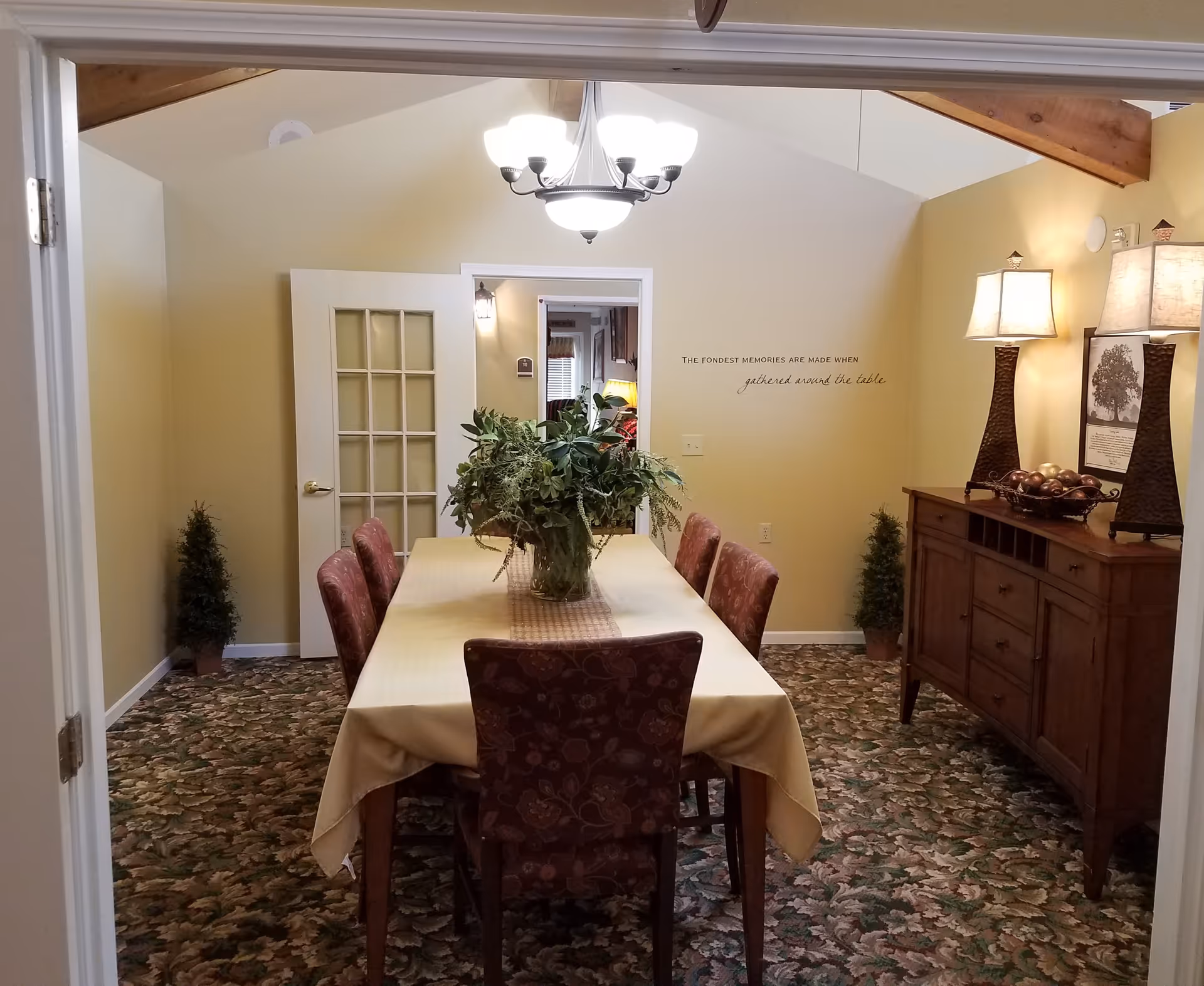 A dining room with a rectangular table covered with a beige tablecloth and six upholstered chairs around it. A large green plant centerpiece is on the table. The room has floral patterned carpet, beige walls, and a wooden sideboard with two lamps and decorative items. A chandelier hangs from the ceiling, and there are two small potted plants in the corners. A wall decal reads 'THE FONDEST MEMORIES ARE MADE WHEN gathered around the table'.