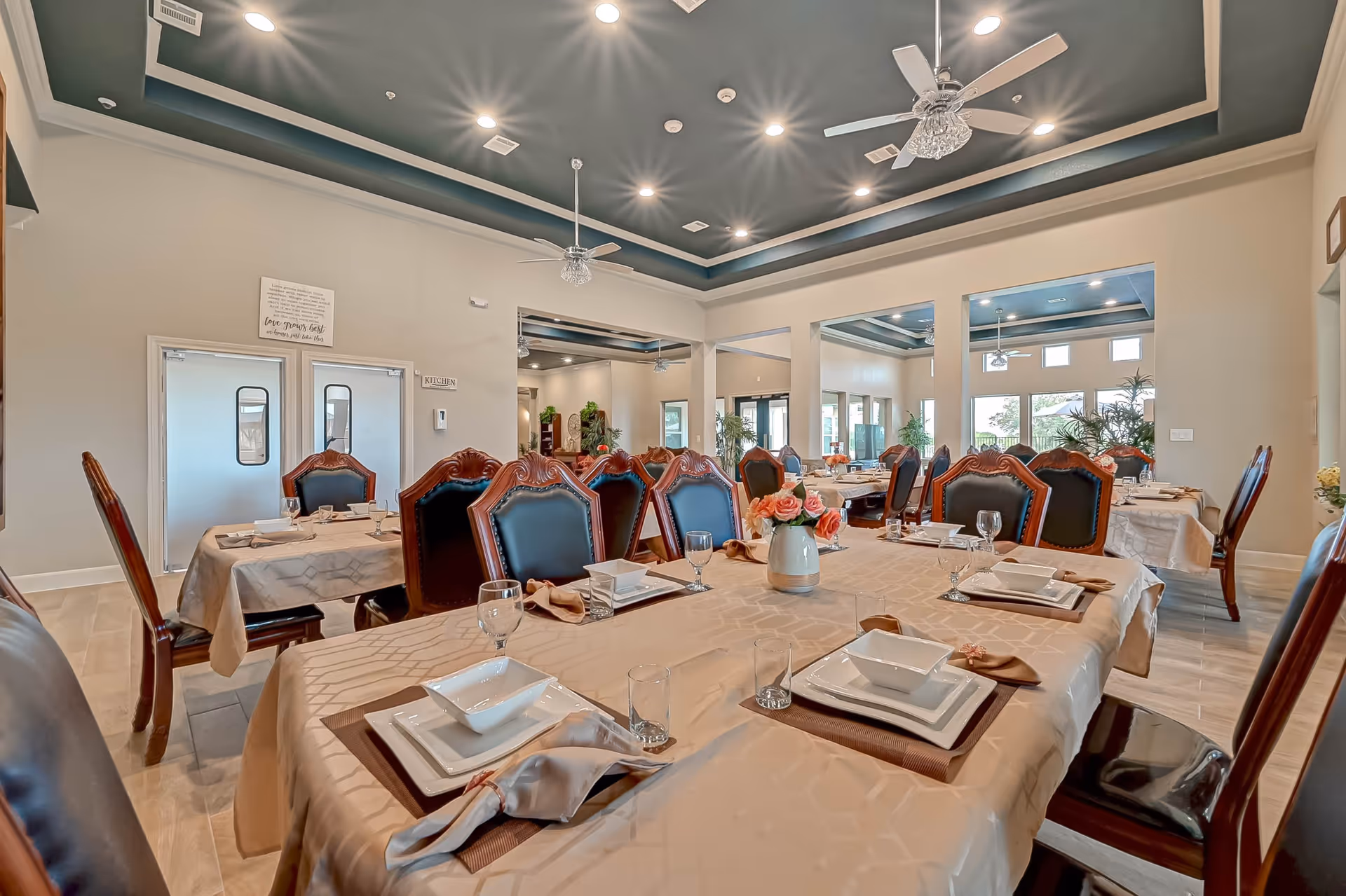 A spacious dining room with multiple tables covered in beige tablecloths, each set with white square plates, bowls, glasses, and beige napkins. The room features elegant wooden chairs with dark leather upholstery, ceiling fans with lights, recessed lighting, and large windows letting in natural light. There are plants and decorative elements in the background, and double doors labeled 'Kitchen' are visible on one wall.