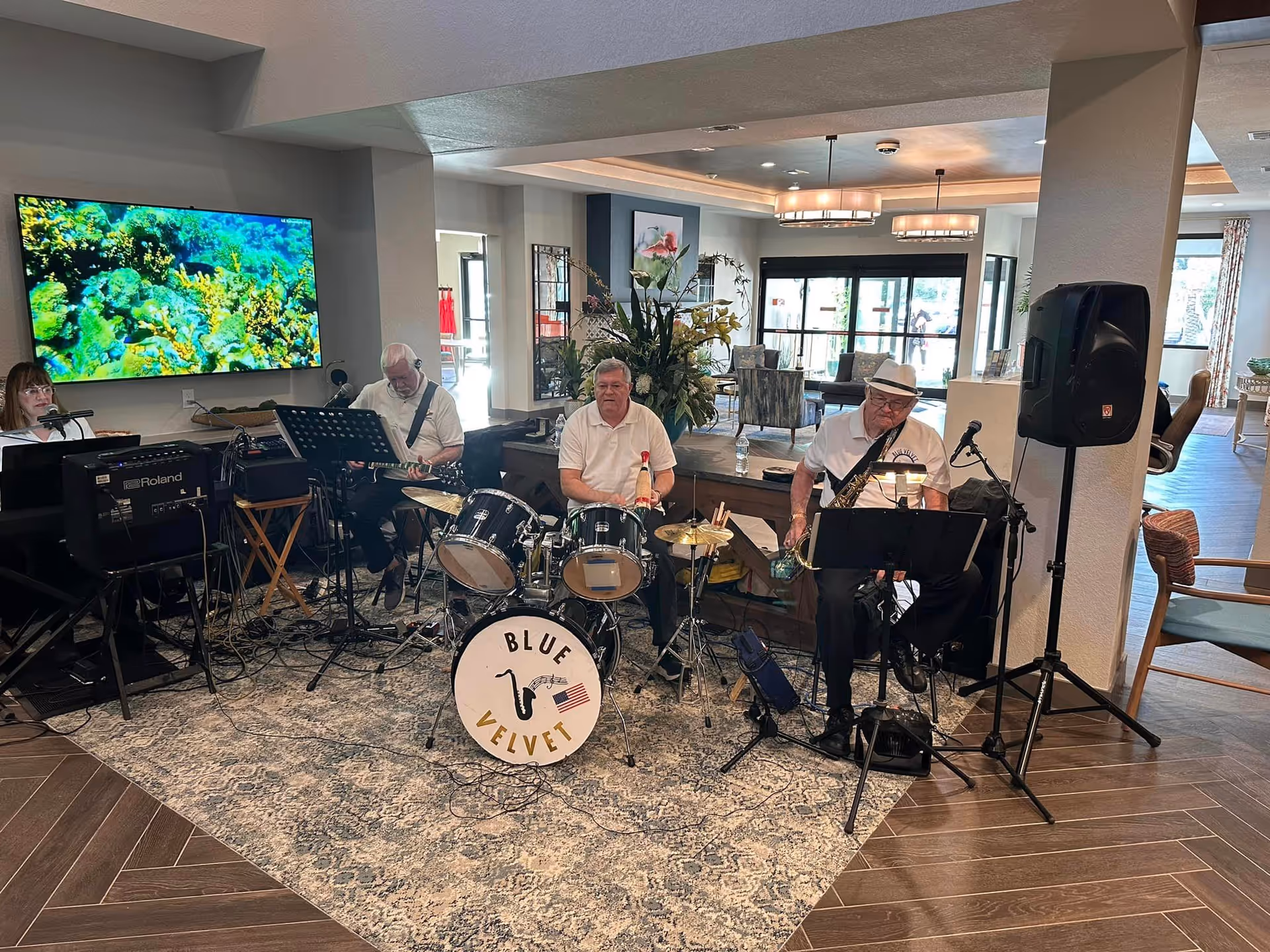 A small band named Blue Velvet performing indoors at The Gallery at Port Orange. The band includes a keyboard player, a guitarist, a drummer, and a saxophonist. They are set up on a patterned rug in a spacious, well-lit common area with modern decor, including a large TV screen displaying an underwater scene, plants, and comfortable seating in the background.