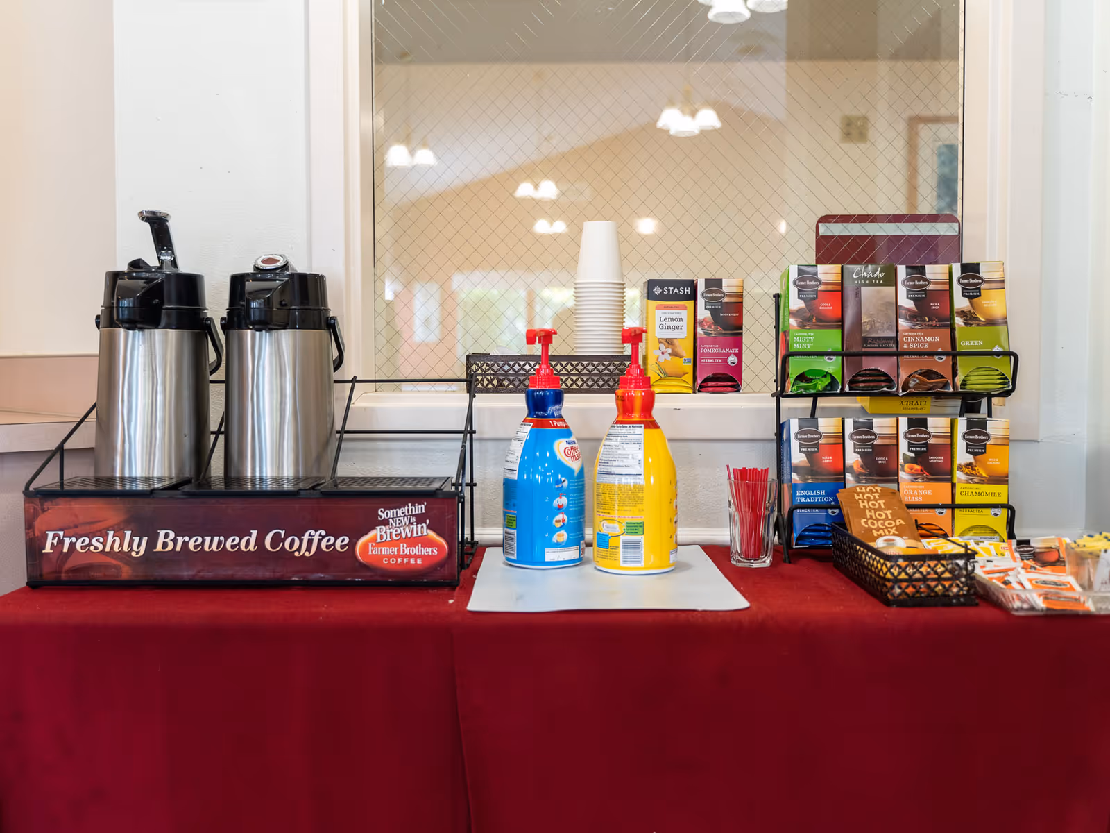 A self-serve coffee and tea station with two airpots, creamer pumps, disposable cups, and a rack of assorted tea bags on a red tablecloth.