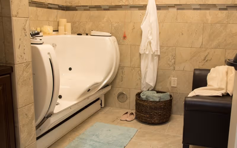 A bathroom with beige tiled walls and floor featuring a white walk-in bathtub with a door, a blue bath mat, a basket with towels, a white bathrobe hanging on the wall, pink slippers on the floor, and a dark brown cushioned chair with a white towel draped over it.