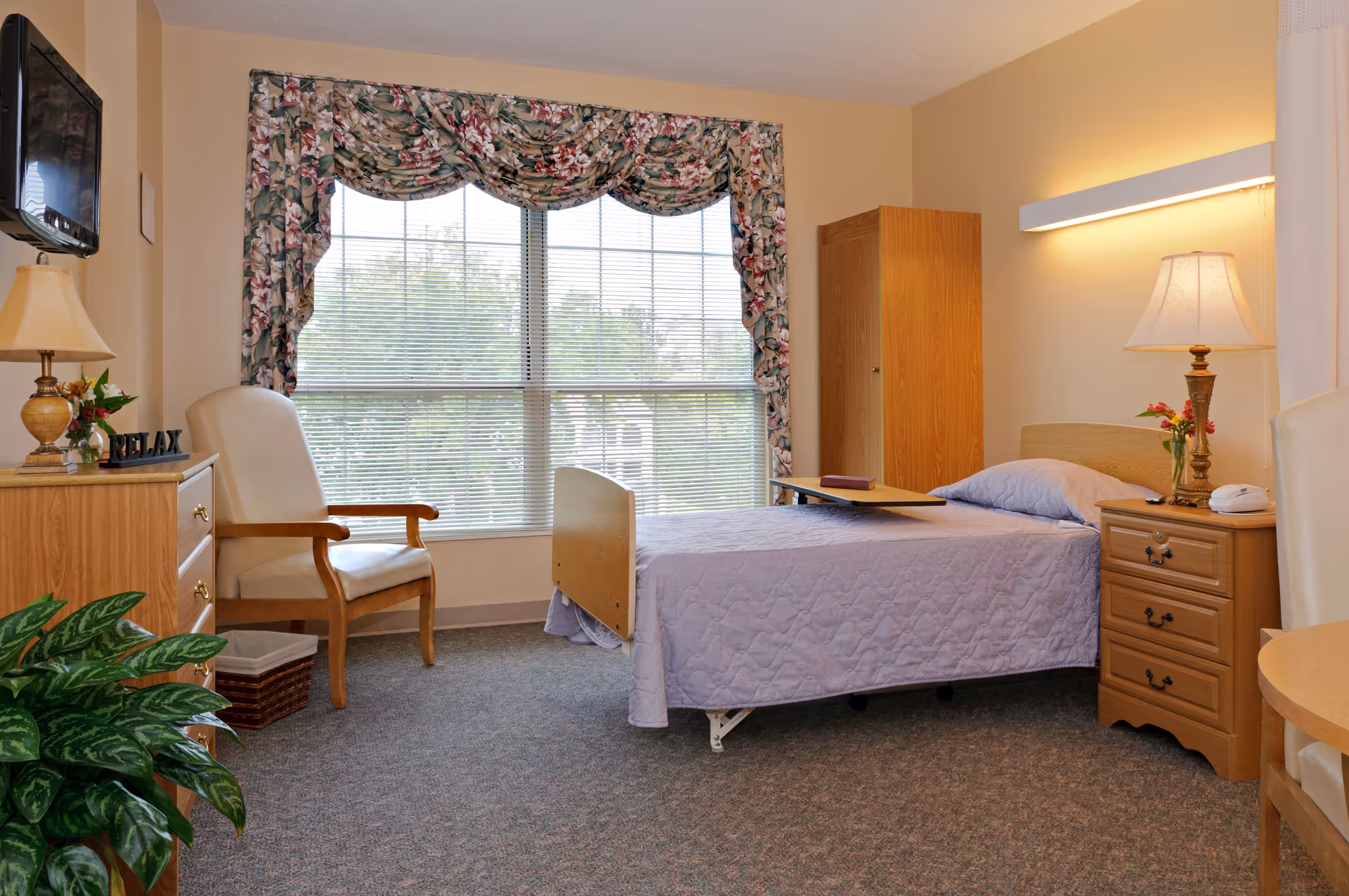 A cozy nursing center bedroom with a single bed covered in a light purple quilt, a wooden nightstand with a lamp and flowers, a wooden wardrobe, a comfortable armchair, a wooden dresser with a lamp and decorative items, a large window with floral curtains, and a green plant in the corner.