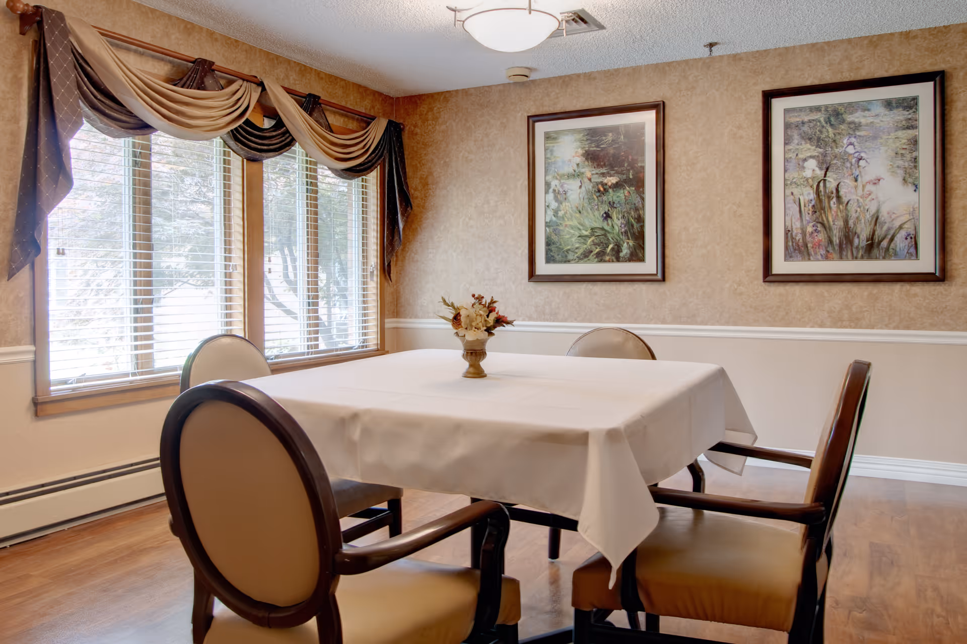 A dining room with a square table covered with a white tablecloth, surrounded by four cushioned chairs with wooden frames. A small floral arrangement is centered on the table. The room has a large window with blinds and decorative drapes, beige walls with white trim, and two framed floral paintings hanging on the wall.