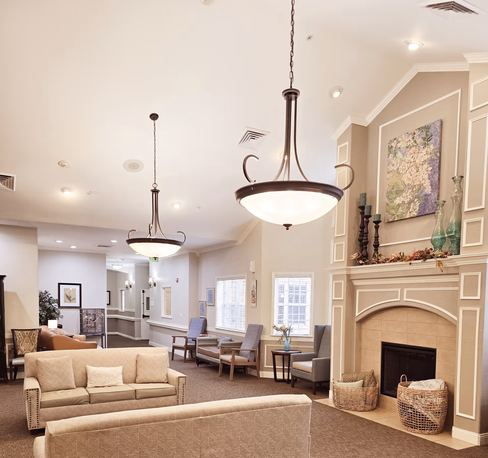 A cozy living room area in a senior living facility with beige sofas, armchairs, a fireplace with decorative vases and candles, a painting above the fireplace, and two large hanging light fixtures. The room has neutral-colored walls and carpeted floors, with windows letting in natural light.