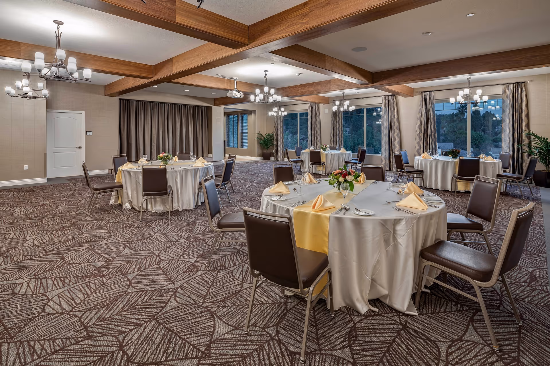 A spacious dining room with round tables covered in white tablecloths and yellow runners, each set with folded napkins, glassware, and floral centerpieces. The room features patterned carpet, wooden ceiling beams, multiple chandeliers, large windows with curtains, and a neutral color palette.