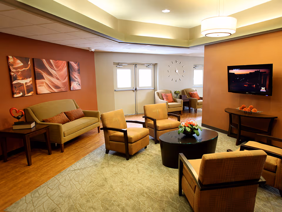 A cozy living room area with a green sofa, four cushioned armchairs arranged around a round black coffee table with a flower arrangement, a wall-mounted flat screen TV, and a wall clock. The room has warm earth-tone walls, wooden flooring, and decorative artwork on the wall.
